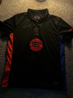 Svart FC Barcelona Nike Match Tröja  - Fet sop man från Nike och FC Barcelona i svart med klubbmärke på bröstet och röd Spotify-logga framtill. Tröjan har röda och blå detaljer på sidorna och korta ärmar. Perfekt för dig som älskar fot-inspirerad stil.