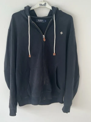 Svart zip-hoodie från Morris - Svart hoodie med dragkedja från Morris. Klassisk modell med vit dragsko i huvan och två stora fickor framtill. Liten broderad logga på bröstet. Tillverkad i mjukt bomullsmaterial för en skön och avslappnad stil.