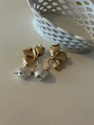 Guldiga örhängen med pärlor och blad - Unika örhängen med guldfärgade blad och oregelbundna vita pärlor. Bladen har en detaljerad struktur och örhängena har en elegant och modern design. Perfekt för dig som vill sticka ut med snygga accessoarer.