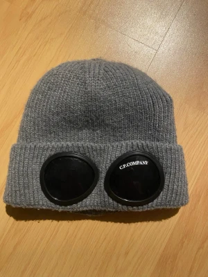 Grå C.P. Company mössa med goggles - Säljer en grå ribbstickad mössa från C.P. Company med ikoniska svarta goggles framtill. Mössan har vikt kant och är tillverkad i ett mjukt, värmande material. Perfekt för dig som vill ha en streetwear-inspirerad look.