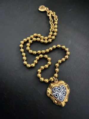 Venturina Paris halsband - Samlarobjekt. Unikt halsband i guldfärgad metall med pärlformade länkar och ett hjärthänge. Hänget har en oregelbunden form och är dekorerat med svart och vitt blommönster. Perfekt för dig som vill sticka ut med accessoarer som känns både trendiga och personliga.
