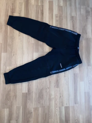 Svarta mjukisbyxor från McKenzie - Säljer ett par svarta mjukisbyxor från McKenzie med resår i midjan och muddar vid bensluten. Byxorna har vita och blå detaljer längs sidorna och en liten logga på benet. Perfekta för chill eller träning.