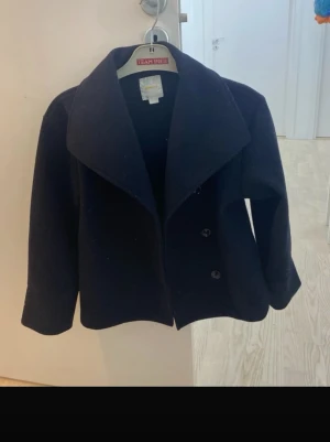 Marinblå skepparkavaj med bred krage - Stilren marinblå skepparkavaj med bred krage och dubbelknäppning framtill. Klassisk rak passform och långa ärmar. Perfekt för dig som vill ha en tidlös och snygg jacka till hösten eller vintern. Materialet ser ut att vara ullblandning.