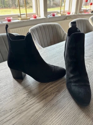 Svarta stövletter i mocka - Snygga svarta stövletter sopassformade i mocka med spetsig tå och bred klack. Skorna har dragflikar bak för enkel påtagning och en stilren siluett som passar till många outfits. Perfekta för dig som gillar en klassisk men trendig look.