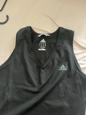 Svart träningslinne från Adidas - Snyggt svart träningslinne från Adidas med v-ringning och diskret logga på bröstet. Tillverkat i ett lätt och stretchigt syntetmaterial som andas, perfekt för gymmet eller löprundan. Ärmlös design för maximal rörelsefrihet.