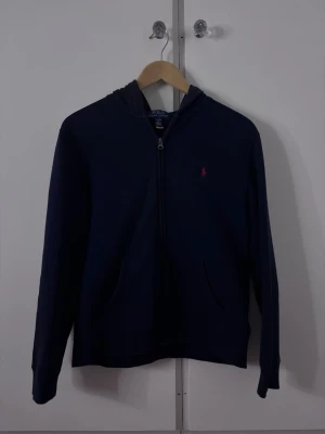 Mörkblå hoodie från Polo Ralph Lauren - Snygg mörkblå hoodie från Polo Ralph Lauren med dragkedja framtill och klassisk huva. Tröjan har två fickor och den ikoniska röda logotypen broderad på bröstet. 