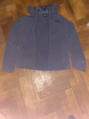 Svart träningsjacka från Under Armour - Svart träningsjacka med huva från Under Armour. Jackan har dragkedja framtill, två sidofickor med dragkedja och en diskret logga på bröstet. Tillverkad i ett lätt och stretchigt material som passar perfekt för träning och sportaktiviteter.