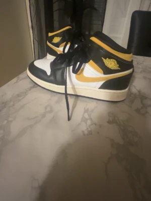 Air Jordan 1 Mid svart/gul/vit - Säljer ett par Air Jordan 1 Mid sneakers i svart, gul och vit. Klassisk high-top modell med svart läder, vita paneler och gula detaljer som swoosh och logga. Skorna har svarta skosnören och en vit sula. Perfekta för dig som vill sticka ut med din stil.