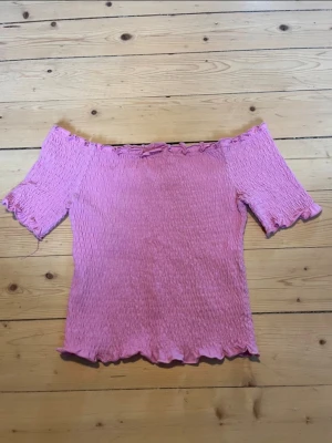 Rosa offshoulder topp i bomull - Söt rosa offshoulder topp med smockad struktur och volangkant. Toppen har korta ärmar och är tillverkad i mjuk bomull med lite stretch för en bekväm passform. Perfekt för en somrig look.