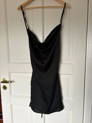 Svart kort klänning från Parisa Amiri - Snygg svart kort klänning från Parisa Amiri med tunna axelband och draperad urringning. Klänningen har snörning i ryggen för en justerbar passform och är tillverkad i ett glansigt material som ger en lyxig känsla.