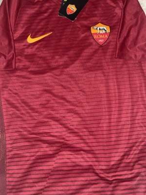 AS Roma matchtröja Nike vinröd 2016 - Säljer en vinröd AS Roma matchtröja från Nike med tunna horisontella ränder och klubbmärke på bröstet. Tröjan har gul Nike-logga och är tillverkad i ett lätt, ventilerande material. Perfekt för fotbollsträning eller att bära som supporter. Den är aldrig använd och köpt i Rom 2016