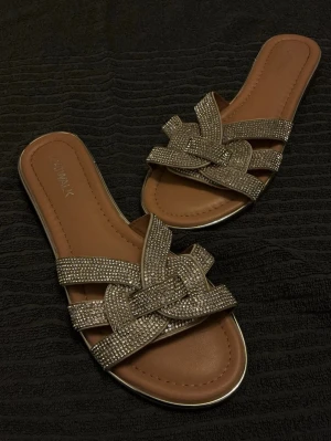 Tofflor med strass - Säljer ett par tofflor från New Walk med flätade remmar täckta av glittrande strass. Sandalerna har öppen tå och platt sula. Passar 38 & 39
