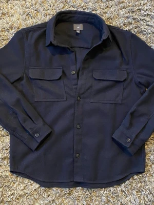 Marinblå overshirt från H&M - Stilren marinblå overshirt från H&M med klassisk krage, två stora bröstfickor med lock och knäppning framtill. Tillverkad i ett mjukt, tjockare tyg som ger en avslappnad look. Perfekt att bära över en t-shirt, skjorta eller hoodie för en trendig lager-på-lager stil. - Skick: 8/10 ✅- Storlek M - Fråga vid minsta fundering!