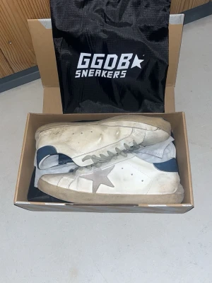 Golden Goose  - Golden Goose sneakers i vitt läder med grå stjärna på sidan och blå häl. Klassisk låg modell med slitna detaljer och beige sula. Skorna har perforeringar på sidan och vita skosnören. Kommer med originalkartong och dustbag. Sulan under är lite paj på båda sidorna en där bak o en där fram men det kan fixas med sko lim❗️❗️❗️