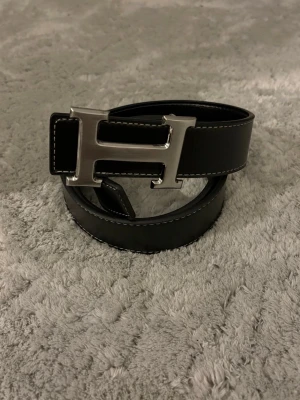 Svart bälte med silver H-spänne  - Snygg svart bälte med stort H-format metallspänne i silver. Bältet har vita kontrastsömmar längs kanterna och är gjort i slätt skinn. Perfekt accessoar för att lyfta din outfit sopphist.