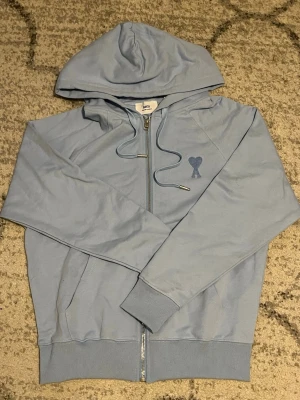 Ljusblå zip hoodie från Ami Paris - Snygg ljusblå hoodie från Ami Paris med dragkedja framtill och huva med dragsko. Tillverkad al bomullsmaterial och har det klassiska Ami-hjärtat broderat på bröstet. Ribbstickade muddar vid ärmslut och nederkant för extra komfort. Helt ny. Storlek S men passar som M