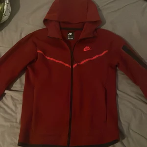 Nike tech hoodie - Säljer en snygg röd Nike tech med dragkedja och huva. Den har en stilren design med Nike-logga på bröstet och en dekorativ linje över bröstet. Perfekt för en sportig look.