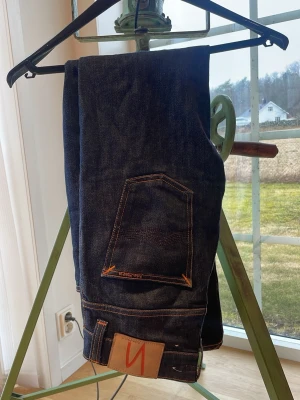 Mörkblå jeans från Nudie Jeans - Säljer ett par mörkblå jeans från Nudie Jeans med klassisk femficksmodell och kontrastsömmar i orange. Jeansen har rak passform och är tillverkade i rå denim för en stilren look. Perfekta för dig som gillar tidlös och clean stil. Jeansen blir snyggare med tiden då vid användning byxans färg/material slits och ger en mer använd/gammal dags look. Strl 29/32.