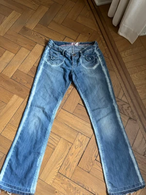 Blå bootcut jeans med blekta kanter och fickor - Säljer ett par blå bootcut jeans med slitningar och ljusa tvättade detaljer. Jeansen har dekorativa sömmar och broderier på fickorna samt råa kanter vid bensluten. Sjukt snygga strl 38-40