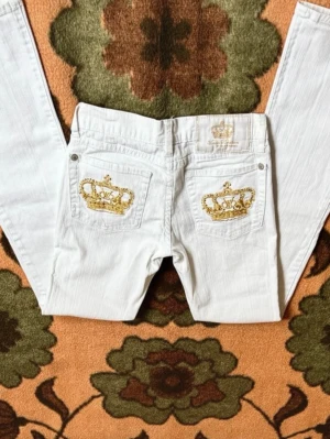 Vita jeans från Victoria Beckham - Säljer ett par vita jeans från Victoria Beckham bootcut med snygga guldbroderade kronor på bakfickorna. Jeansen har klassisk femficksdesign och patch med logga bak i midjan. Perfekta för dig som vill sticka ut med exklusiva detaljer. Om du har några frågor är det bara att höra av sig! Säljer då det ej passade