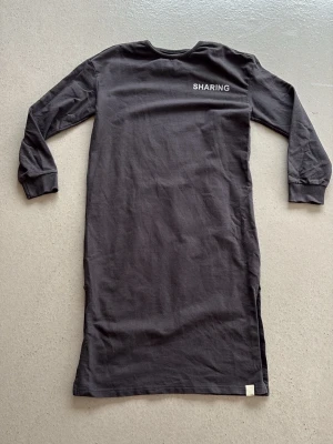 Svart långärmad klänning I Dig Denim - En svart långärmad klänning från I Dig Denim med rund halsringning och trycket 'SHARING' framtill. Klänningen har en rak passform och är tillverkad i mjuk bomull, perfekt för en avslappnad och enkel stil.