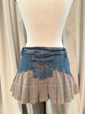 Y2K jeans pleated kjol  - Säljer en unik kortkjol med jeans upptill och rutigt, plisserat tyg nedtill. Kjolen har tre dekorativa knappar framtill och bälteshällor. Perfekt för dig som gillar Y2K med en mix av denim och klassiskt mönster.