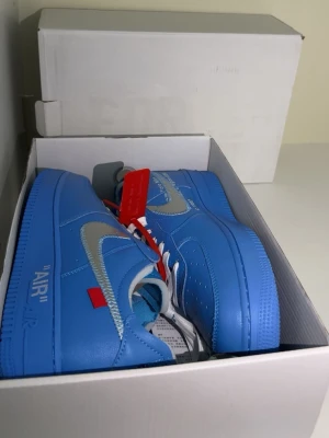 Nike x Off-White Air Force 1 blå - Säljer ett par Nike x Off-White Air Force 1 sneakers i en riktigt snygg blå färg med silver swoosh och vita skosnören. Skorna har Off-White detaljer som röd tag och text på sidan. Tillverkade i läder med klassisk Air Force 1 sula. Perfekta för dig som gillar streetwear.