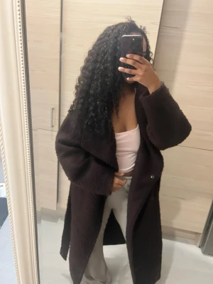 Mörkbrun teddykappa Gina  - Säljer en lång mörkbrun teddyrock med bred krage och stora slag. Rocken har en avslappnad passform och är gjord i ett mjukt, fluffigt material som ger en trendig look. Perfekt för att hålla sig varm under kyliga dagar.