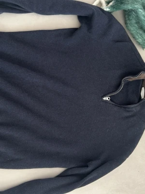 Mörkblå stickad tröja med halvzip - En stilren mörkblå stickad tröja med halvzip och hög krage. Tröjan har långa ärmar och ribbade muddar vid ärmslut och nederkant. Perfekt för lager-på-lager och enkel att matcha med jeans eller joggers.