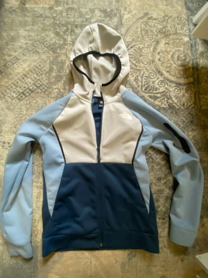Blå och grå hoodie från The North Face - Snygg hoodie från The North Face i blått och ljusgrått med dragkedja framtill och huva. Hoodien har svarta detaljer och en logga på bröstet. Tillverkad i mjukt och bekvämt material, perfekt för chill dagar eller när du vill ha en sportig look.