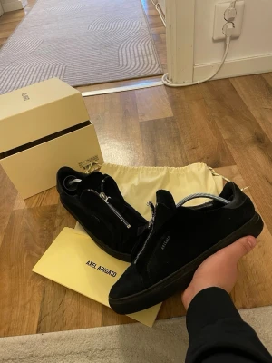 Svarta sneakers från Axel Arigato zip 90 - Stilrena svarta sneakers från Axel Arigato i mocka med coola silverfärgade dragkedjor istället för vanliga snören. Låg modell med svart sula och diskret logga på sidan. Perfekta för dig som vill ha en clean och modern look.