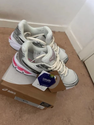 Vita och silver Asics sneakers - Snygga Asics sneakers i vitt och silver med rosa detaljer. Skorna har mesh-ovandel för bra andningsförmåga och Ortholite-innersula för extra komfort. Klassisk låg modell med vita snören och robust sula, perfekta för en sportig och trendig look. Kan sänka priset vid snabb affär/ helt nya köpte fel storlek 