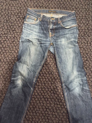 Blå jeans från Nudie Jeans - Säljer ett par klassiska blå jeans från Nudie Jeans med snygga slitningar och kontrastsömmar. Jeansen har rak passform, fem fickor och detaljer i orange. Tillverkade i denim av bomull.