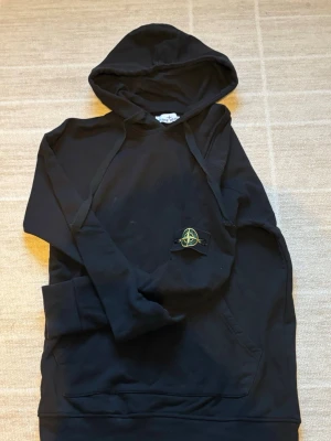 [STONE ISLAND] hoodie  - Snygg svart hoodie från Stone Island med klassisk logotyp-patch på ärmen. Hoodien har huva med dragsko och en stor magficka framtill. Tillverkad i mjukt bomullsmaterial som känns skönt mot huden. Perfekt för en avslappnad streetwear-look. Äkta har taggarna har ej ändvänt därför tänk sälja den 