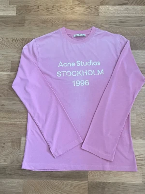 Rosa långärmad topp Acne Studios - Långärmad rosa topp från Acne Studios med tryck i vitt framtill där det står 'Acne Studios STOCKHOLM 1996'. Tillverkad i mjuk bomull och har en klassisk rund halsringning. Perfekt för en avslappnad och trendig look.