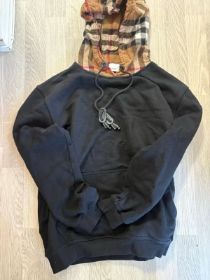 Svart hoodie med Burberry-rutig huva - Svart hoodie från Burberry med klassisk rutig huva i beige, svart, vitt och rött. Hoodien har dragsko vid huvan och en avslappnad passform. Perfekt för dig som gillar streetwear och vill sticka ut med en ikonisk detalj.  Säljer den helt enkelt för har gått ner i vikt och den passar ej mig mer. Helt ny då haft på mig den bara 2-3 gånger 