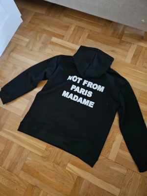 Svart hoodie Not From Paris Madame - Svart hoodie från Drole de Monsieur med vit text 'NOT FROM PARIS MADAME' tryckt på både bröstet och ryggen. Klassisk passform med huva och dragsko samt känguruficka framtill. Perfekt för dig som gillar streetwear och tydliga statements. Storlek M men sitter som ett S