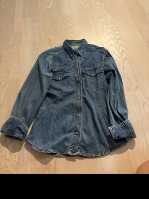 Blå jeansjacka från Girls Casual Wear - Snygg blå jeansjacka från Girls Casual Wear med klassisk krage, två bröstfickor med lock och knappar framtill. Jackan har långa ärmar och en avslappnad passform som ger en cool vintagekänsla. Perfekt att slänga över en hoodie eller t-shirt.