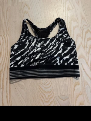Svart och vit sport-bh med mönster - Snygg sport-bh i svart och vitt med abstrakt mönster och bred elastisk randig resår nedtill. Racerback-modell för bra stöd och rörelsefrihet. Perfekt för träning och sportaktiviteter. Tillverkad i stretchigt funktionsmaterial.