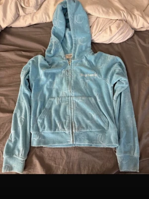 Blå hoodie från Juicy Couture - Säljer en ljusblå hoodie från Juicy Couture med dragkedja framtill och stor ficka. Hoodien har JC-loggor i relief över hela tyget och en klassisk huva. Perfekt för en chill och trendig look.