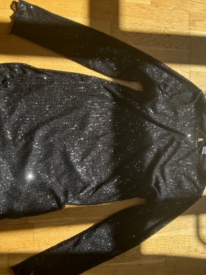 Svart glittrig långärmad klänning - Snygg svart klänning med långa ärmar och glittriga paljettdetaljer som fångar ljuset. Perfekt för dig som vill sticka ut och glänsa. Klänningen har en kroppsnära passform och är i kort modell.