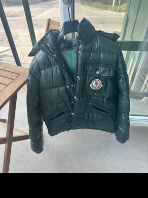 Mörkgrön dunjacka från Moncler - Snygg mörkgrön dunjacka från Moncler med glansig finish, stor huva och Moncler-logga på bröstet. Jackan har en bröstficka med knapp, två sidofickor och dragkedja med tryckknappar. Perfekt för kalla vinterdagar och riktigt trendig look.