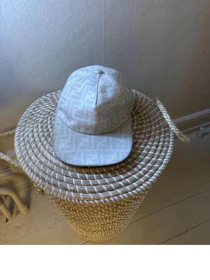 Beige Fendi keps med FF-mönster - Säljer en beige keps från Fendi med det klassiska FF-logomönstret över hela kepsen. Kepsen har böjd skärm och justerbar rem bak. Materialet känns lyxigt och kepsen har svart insida. Perfekt för dig som gillar exklusiva accessoarer.