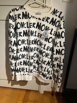 Vit Moncler sweatshirt med logotypmönster - Säljer en vit sweatshirt från Moncler med svart, grafiskt logotypmönster över hela tröjan. Rund halsringning och långa ärmar. Tröjan har en Moncler-patch på bröstet och är tillverkad i mjukt material för en avslappnad look.
