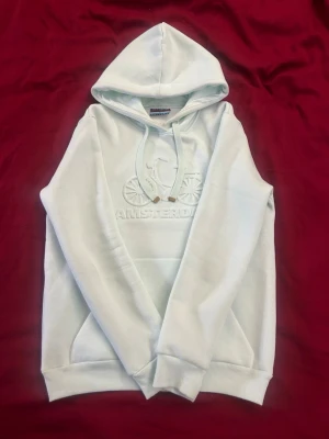 Vit hoodie från Kist Design - Snygg vit hoodie från Kist Design med broderad cykel och texten 'Amsterdam' på bröstet. Hoodien har dragsko i huvan, magficka och ribbade muddar. Tillverkad i mjukt material som känns skönt mot huden. Perfekt för en avslappnad streetwear-look.