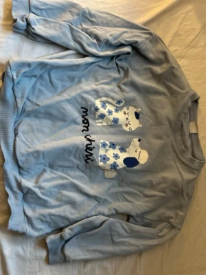 Blå sweatshirt med katt och hund Lindex för barn  - Söt ljusblå sweatshirt från Lindex med tryck av en katt och en hund i blått och vitt samt texten 'mon chéri' framtill. Tröjan har ribbade muddar vid ärmslut och nederkant. Perfekt för dig som gillar gulliga motiv och mjuka tröjor.