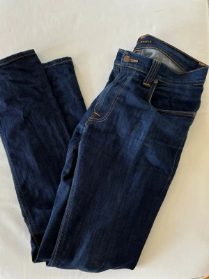 Mörkblå jeans från Nudie Jeans - Säljer ett par mörkblå jeans från Nudie i ett väldigt bra skick. Jeansen har raka ben och snygga broderade detaljer på bakfickorna. 