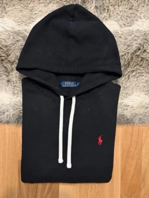  Polo Ralph Lauren Hoodie - Klassisk svart hoodie från Polo Ralph Lauren med vit dragsko och röd broderad logga på bröstet. Hoodien har en stor huva och magficka framtill. Tillverkad i mjukt bomullsmaterial som känns skönt mot huden. Perfekt för en avslappnad och stilren look.