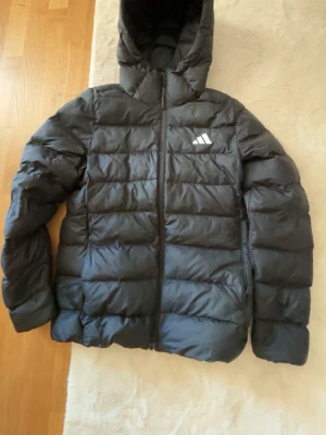 Svart dunjacka från Adidas med huva - Snygg svart dunjacka från Adidas med huva och dragkedja framtill. Jackan har ett diskret Adidas-logotyp på bröstet och quiltad design som håller värmen. Perfekt för kalla vinterdagar och riktigt skön att bära.