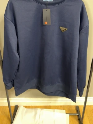 Mörkblå sweatshirt från Prada - Stilren mörkblå sweatshirt från Prada med klassisk rund halsringning och lång ärm. Tröjan har Pradas ikoniska metallogo på bröstet och är tillverkad i mjukt bomullsmaterial. Perfekt för en clean och exklusiv look.